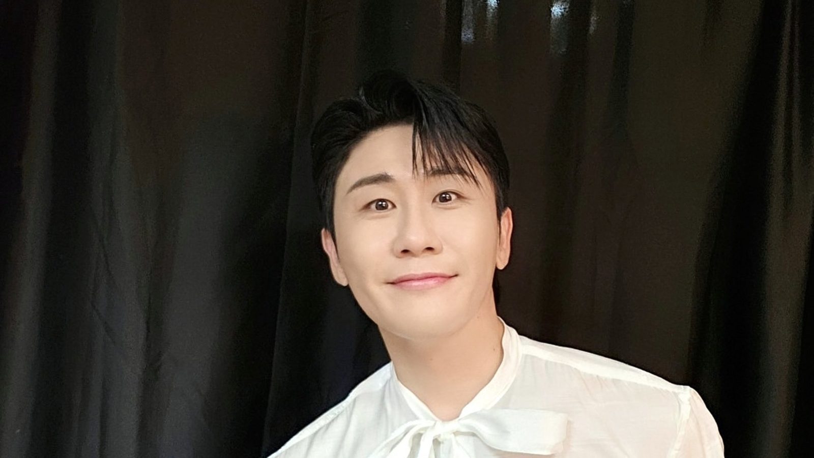 영탁이 하얀 리본 블라우스 차림으로 검은 커튼 앞에서 정면을 보며 은은하게 웃는 모습