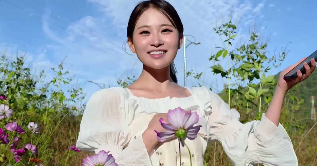 푸른 하늘 아래 코스모스 들판에서 한 손에 휴대전화를 들고 환하게 웃는 김다현