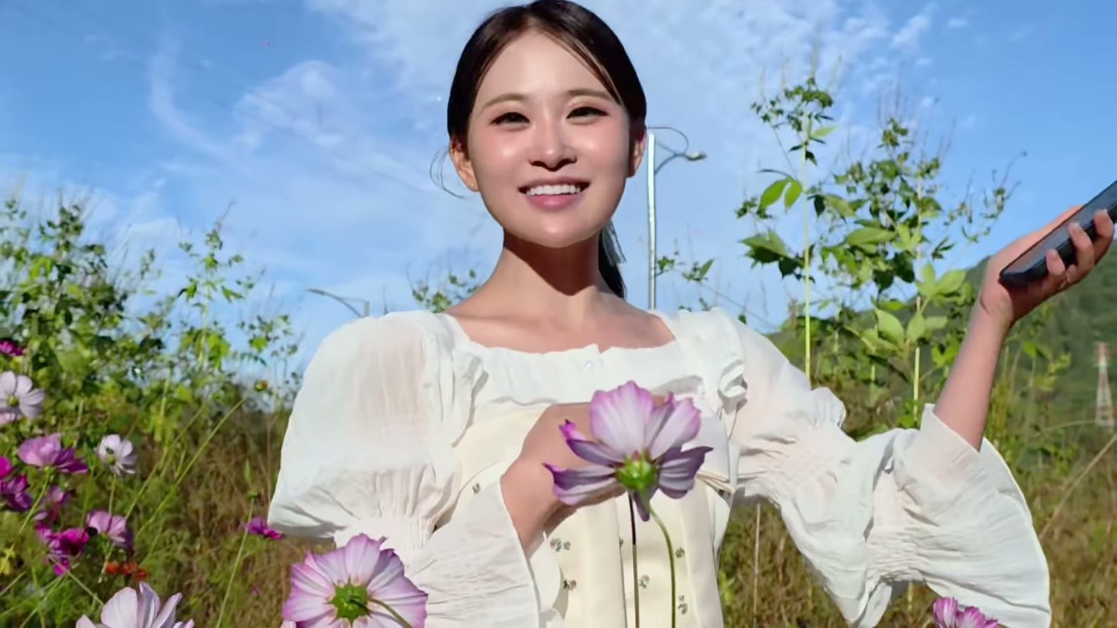 푸른 하늘 아래 코스모스 들판에서 한 손에 휴대전화를 들고 환하게 웃는 김다현