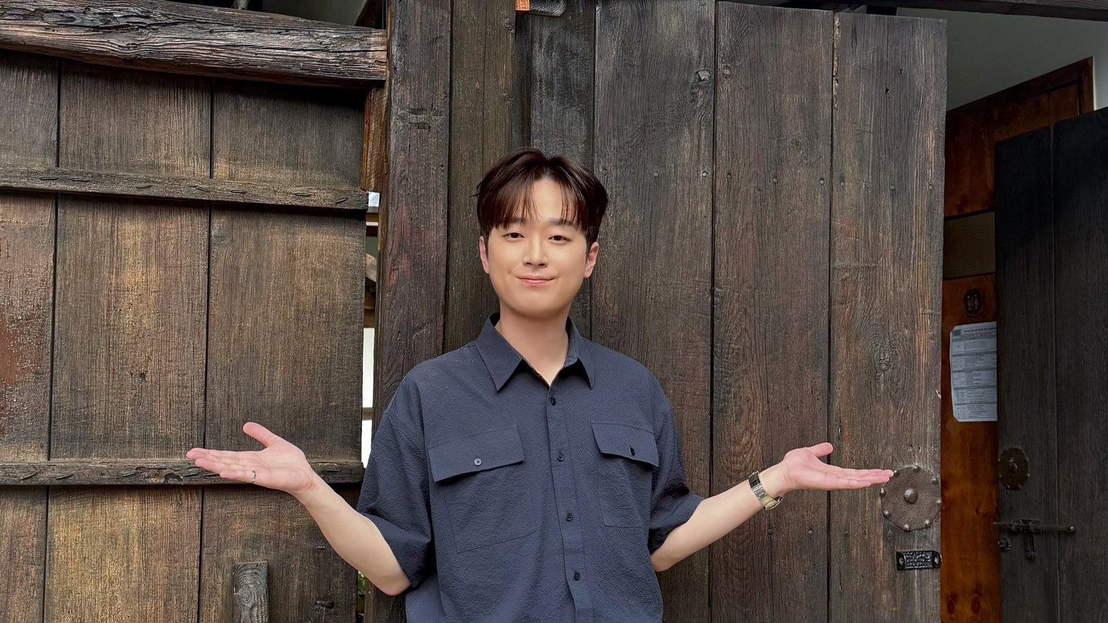 남색 셔츠에 청바지 차림의 이찬원이 오래된 나무 문 앞에서 두 팔을 펼치고 웃는 모습
