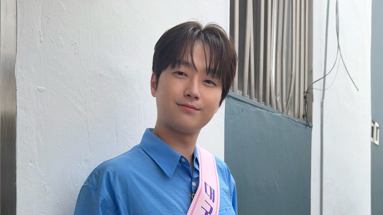 파란 셔츠에 어깨띠를 두르고 흰 벽 앞에서 미소 짓는 이찬원