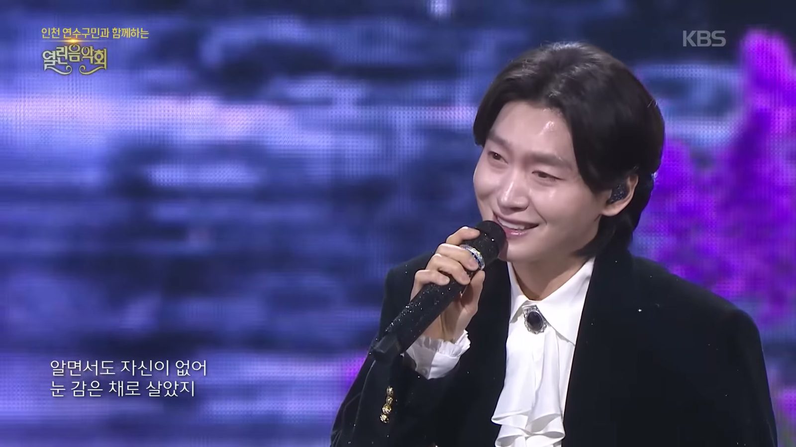 검은 벨벳 재킷에 흰 프릴 셔츠 차림으로 마이크를 쥐고 환하게 웃으며 노래하는 김용빈