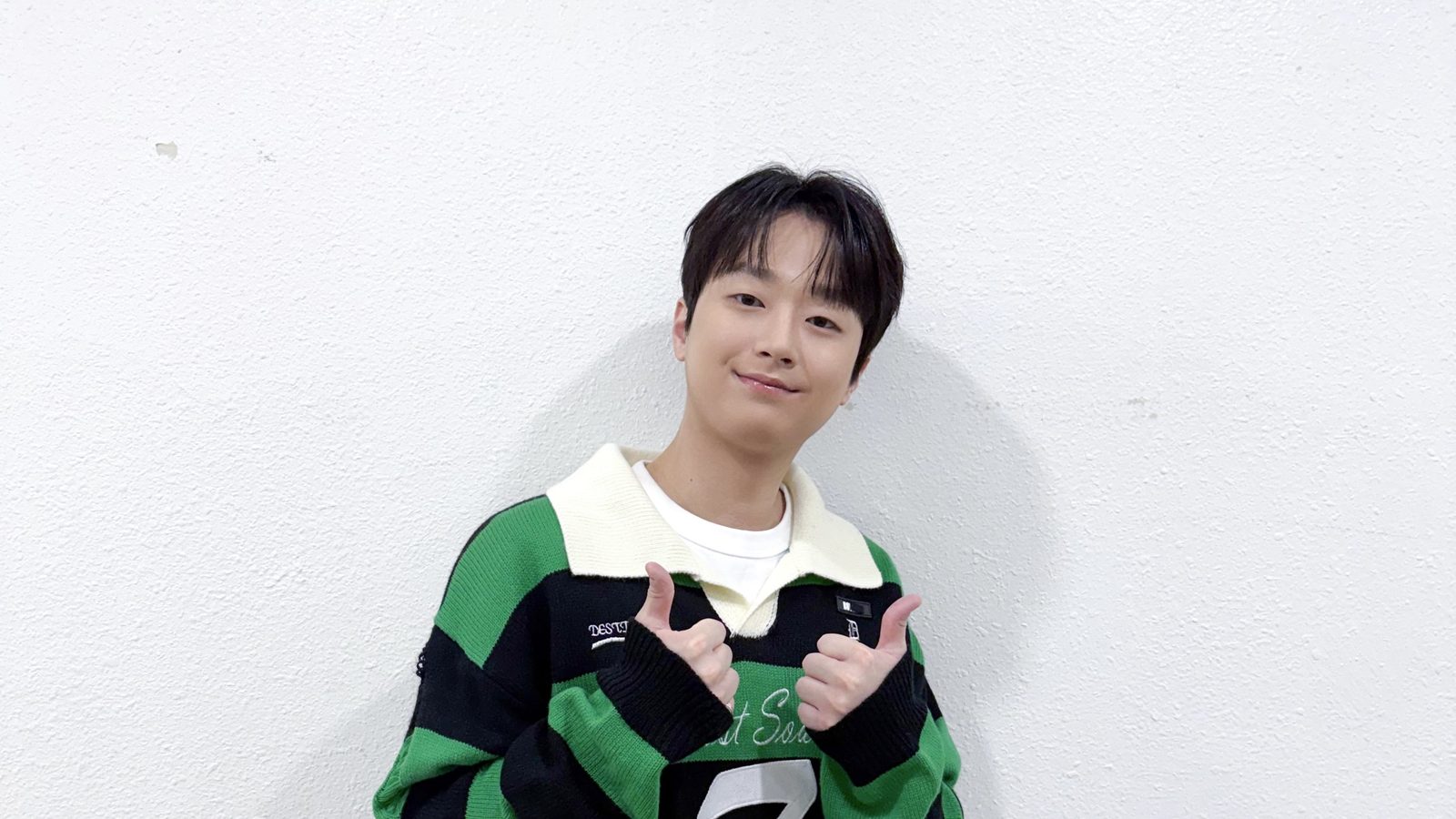 흰 벽 앞에서 초록 줄무늬 상의를 입고 양손 엄지를 들어 보이며 웃는 이찬원